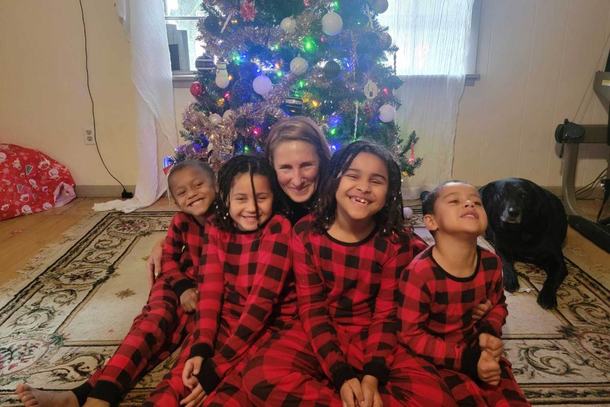 A family of five pose in front of a Christmas tree while wearing matching red and black pajamas as a black dog looks on from behind them