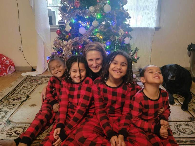 A family of five pose in front of a Christmas tree while wearing matching red and black pajamas as a black dog looks on from behind them
