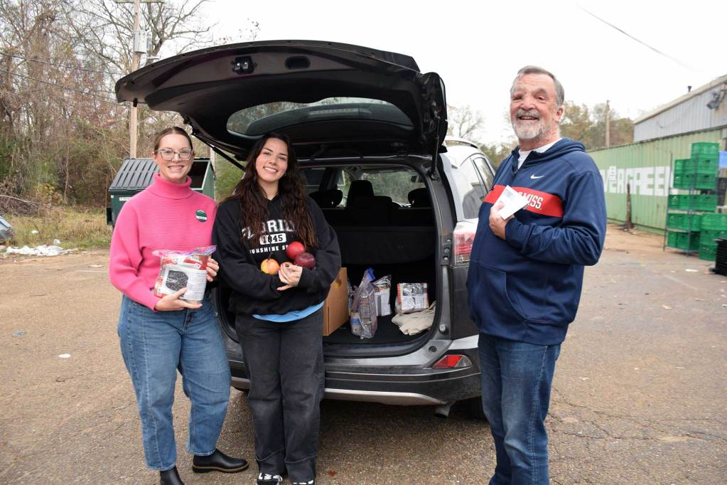 A man and two women pose behind a grey vehicle's open trunk