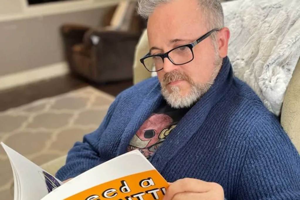 A man sits in a living room chair and reads the book 'I Need A New Butt'