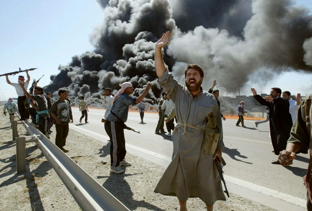 A man holding his hand in the air. A group of people are behind him. Behind the group, a massive plume of gray smoke flows from the ground.