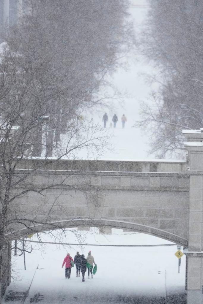 A bridge crosses the snowy view, and small groups of people are walking down the snow covered road beneath it