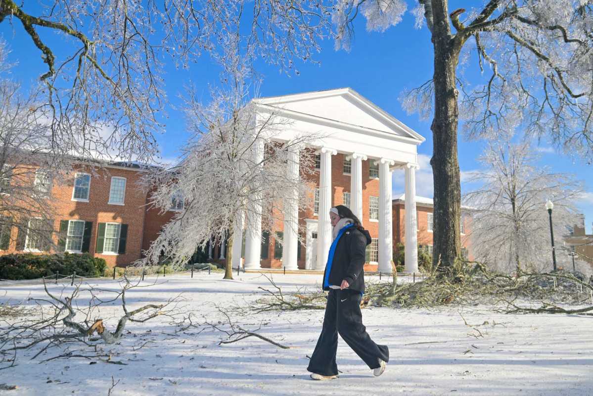 A person in warm clothes walks across a snowy campus, icy fallen branches visible on the grounds