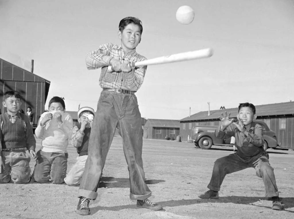 A black-and-white photo of a young boy swinging a baseball bat at a ball. Another young boy stands near him ready to catch the ball.