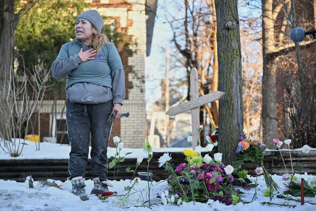 A crying woman stands next to a makeshift memorial with a small wooden cross and decorative flowers.