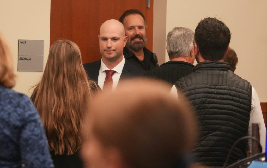 A bald man wearing a suit entering a courtroom with several people in front of him.
