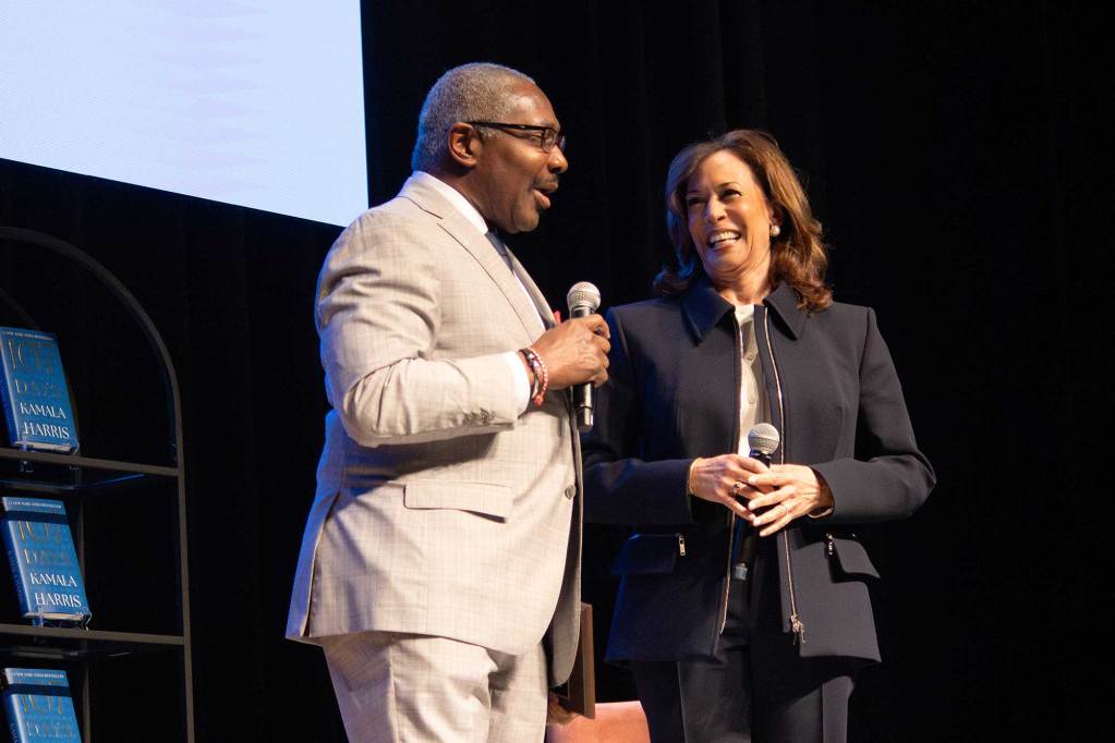 John Horhn in a light colored suit, and Kamala Harris in a dark colored suit speak together on stage