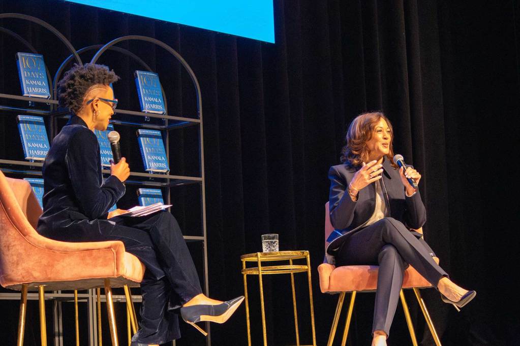 Rita Brent and Kamala Harris speak on stage, seated in pink chairs and backed by shelves holding Harris's new book 107 Days