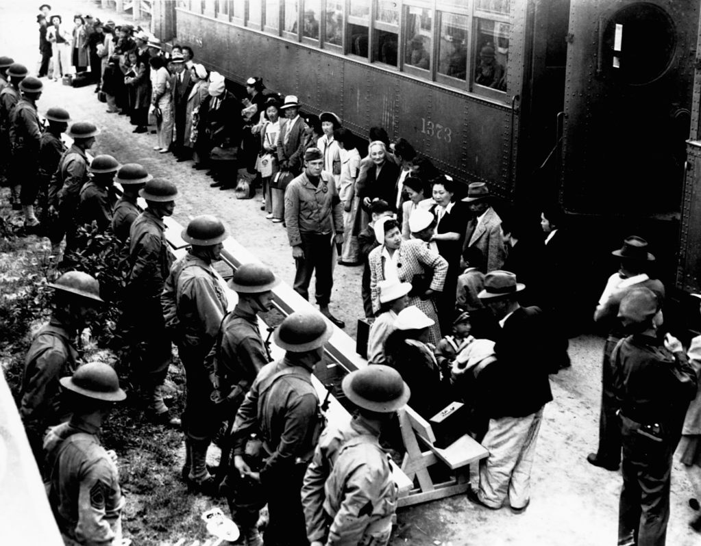 A black-and-white photo of a group of people lined up near a train. In front of them are several soldiers in a line.