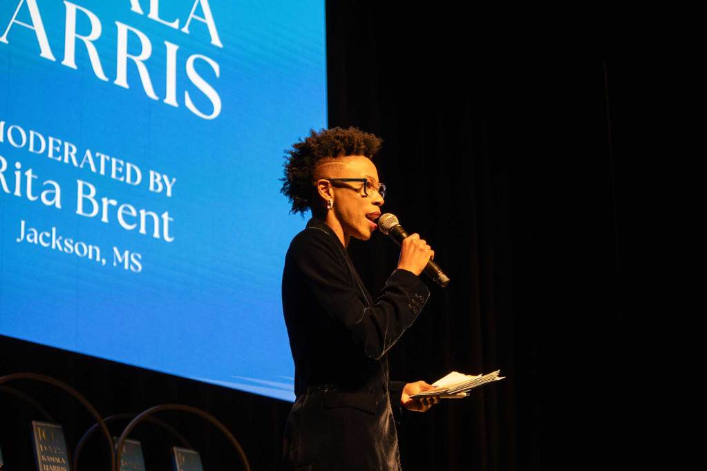 Rita Brent, in a black suit, speaks on stage