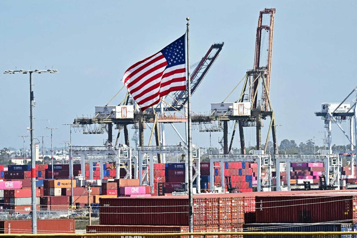 An American flag with a shipping port in the background featuring tall shipping cranes.