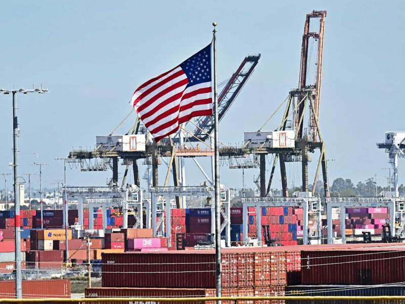 An American flag with a shipping port in the background featuring tall shipping cranes.