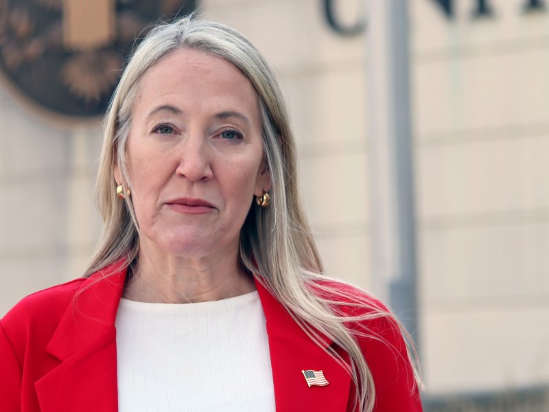 A woman with long blonde hair wearing a red jacket with a US Flag lapel pin stands outside of a federal court building