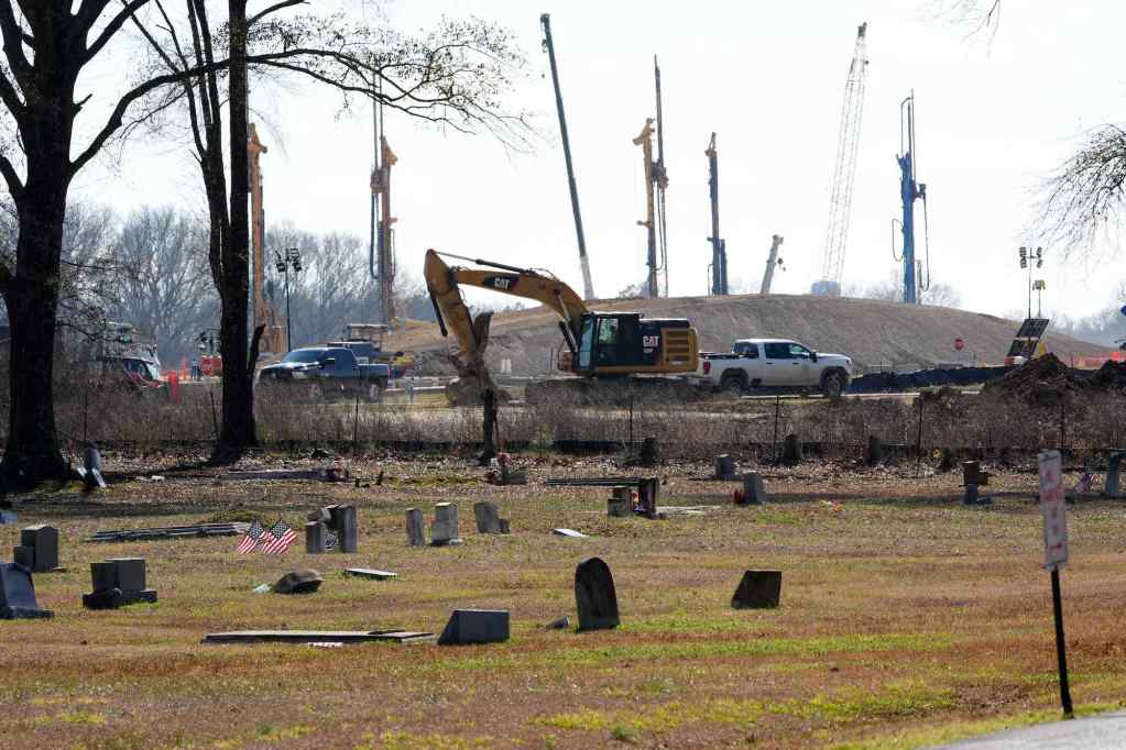 A small cemetery by the road sits in the foreground as large construction equipment works in field right beside it