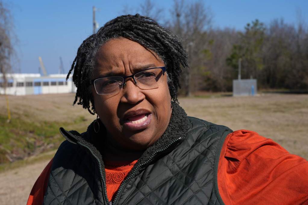 A woman in an orange and black top speaks outside, a data center seen in the distance behind her