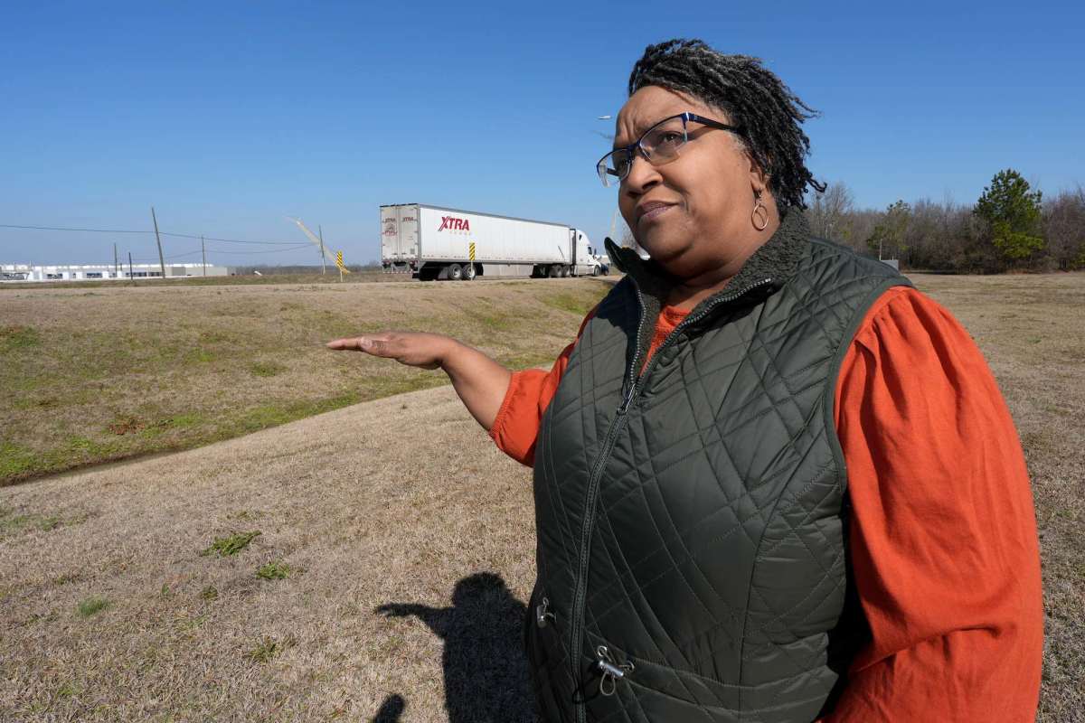 A woman in an orange and black top talks in a field, hand gesturing towards the road