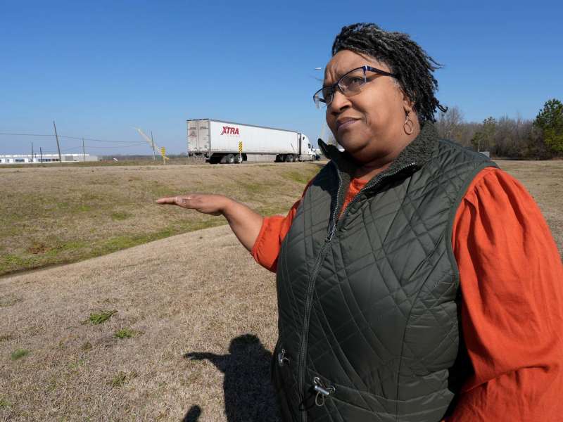 A woman in an orange and black top talks in a field, hand gesturing towards the road