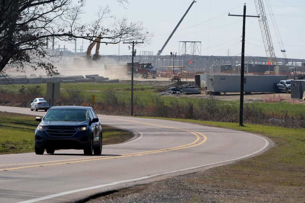 Cars drive down a winding road beside a large active construction site