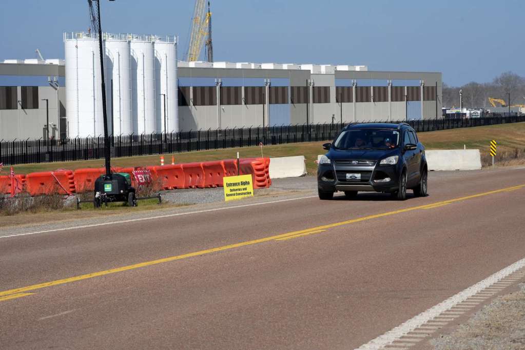 A car drives past a grey blue and white datacenter with orange barriers at the entrance