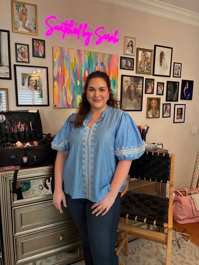 A woman wearing a blue shirt and blue jeans standing. Behind her on the wall are several framed photos and art pieces.