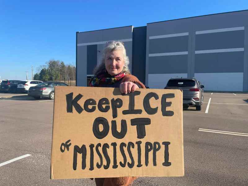 a woman holds a cardboard sign that says "Keep ICE out of Mississippi" while standing in front of a gray warehouse