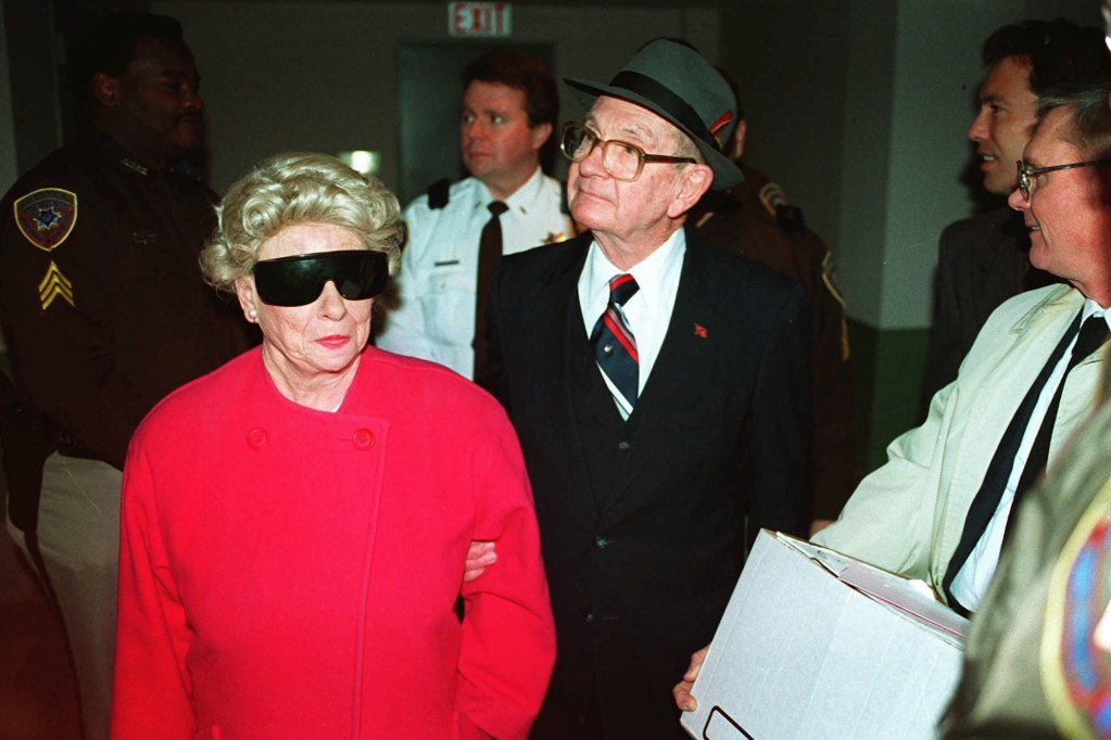 an old white man in a green bowler hat with a red feather in it walks while holding onto the arm of an old white woman with blond hair and large sunglasses