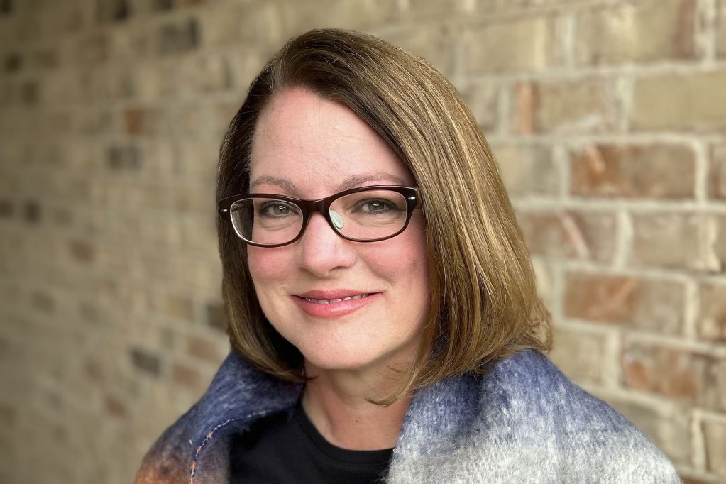 A closeup of Courtney Lipscomb in glasses, smiling in front of a brown brick wall