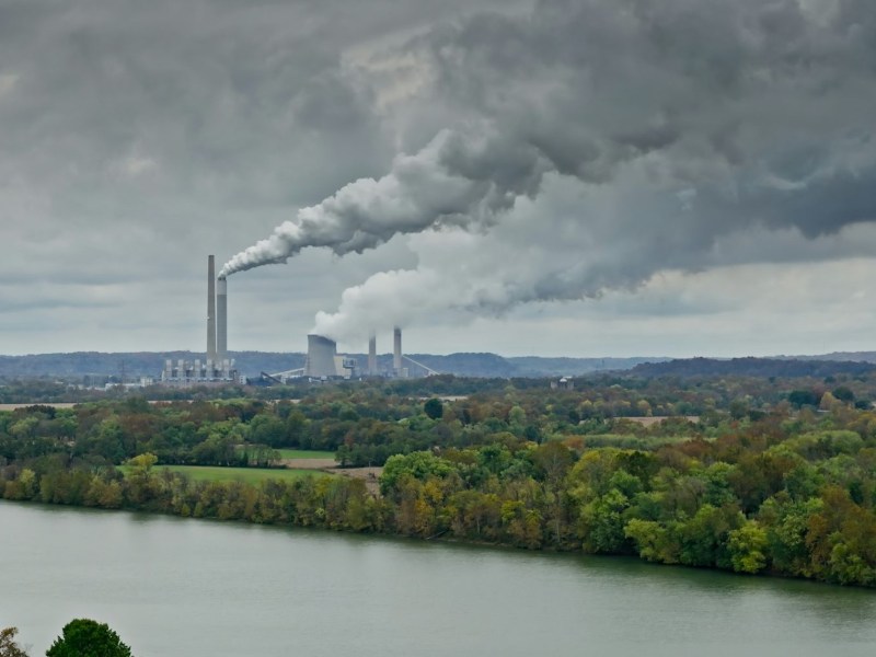 A factory in the background of a forested area with a river. Smoke billows from the factory’s smokestacks.