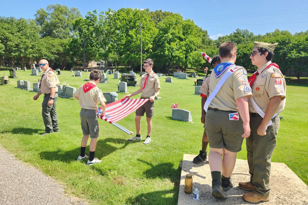 Several boys wearing scout uniforms in a cemetery. Two scouts are folding an American flag.