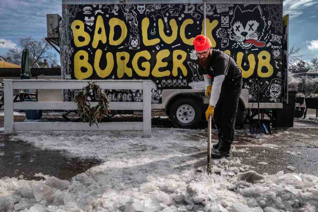 A man shovels snow and ice outside of a black foodtruck labeled 'Bad Luck Burger Club' in handwritten yellow letters