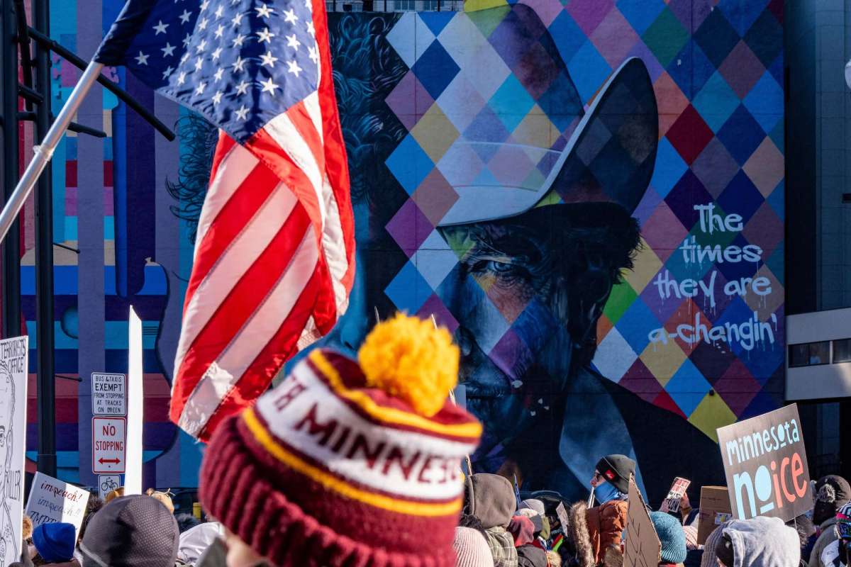 An American flag is held prominently during an ICE protest. In the background is a colorful mural of Boy Dylan with the inscription, “The times they are a-changin.”