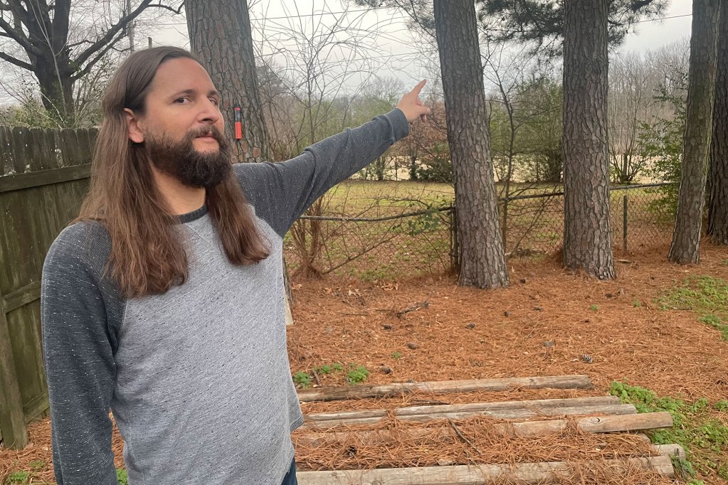 A man in a grey shirt looks at the camera but points behind him, past his back yard