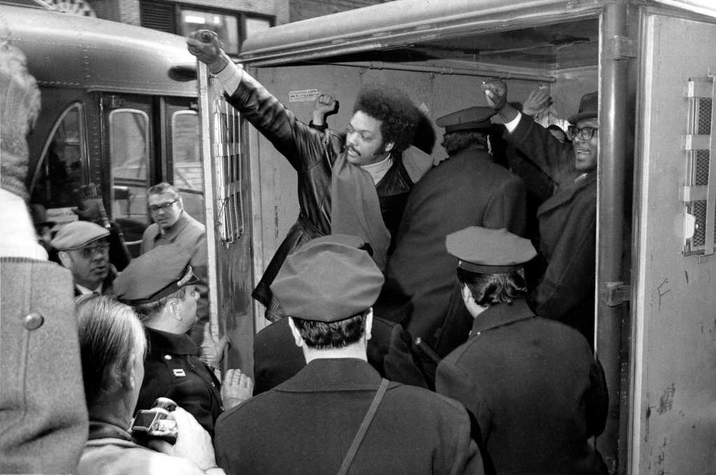 An old black and white photo of Jesse Jackson putting a fist in the air from the back of a police van