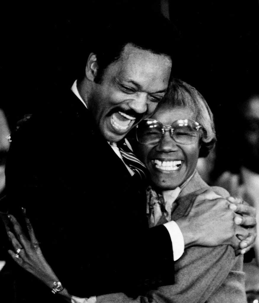 An old black and white photo of Jesse Jackson hugging Shirley Chisholm as they smile
