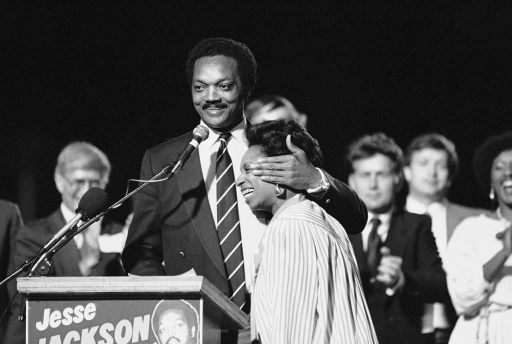 An old black-and-white photo of Jesse Jackson standing at a podium and hugging his wife, Jacqueline.