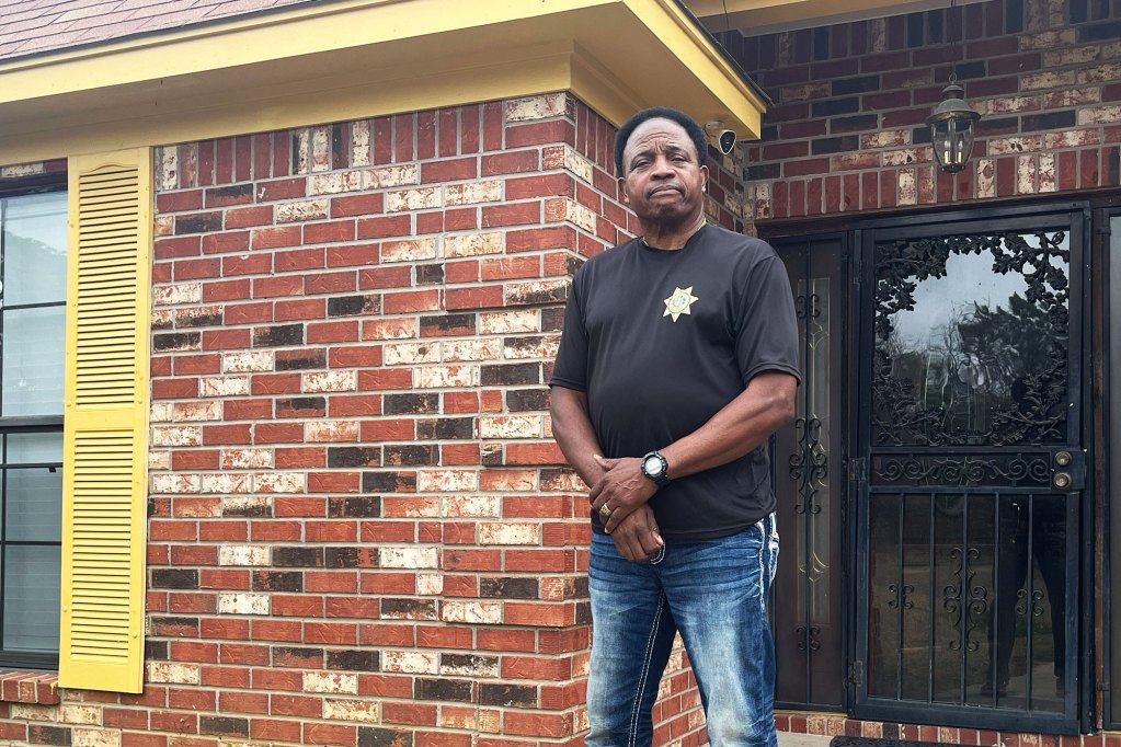 A man wearing a black tshirt with a sheriff badge icon on the chest stands outside of a red brick home with yellow trim
