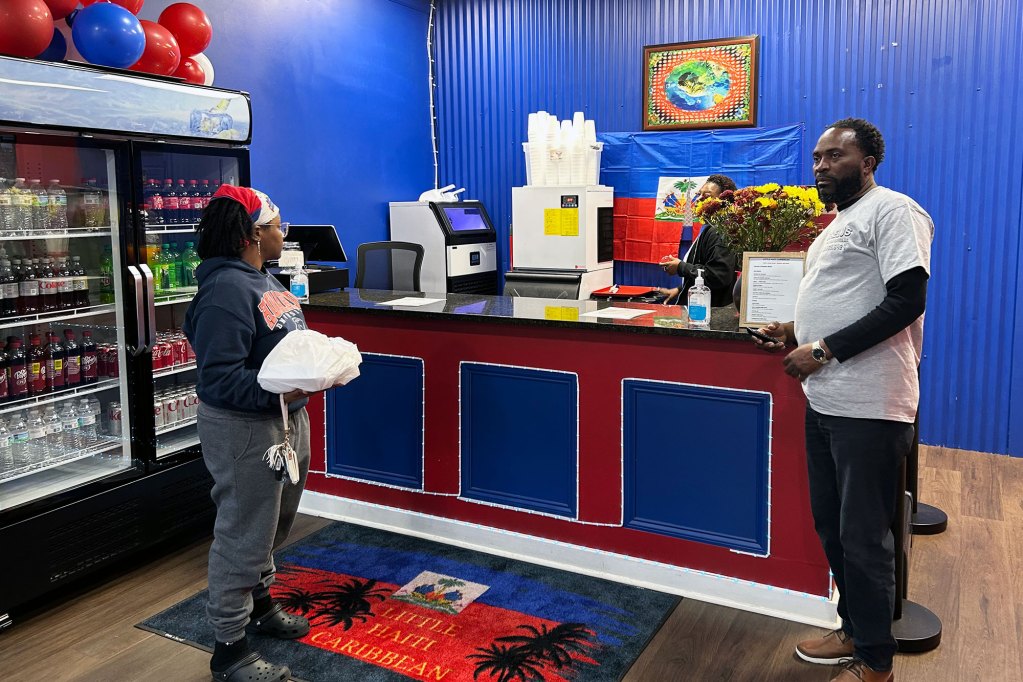 An owner of a restaurant labeled 'Little Haiti Caribbean' speaks to a customer at the counter