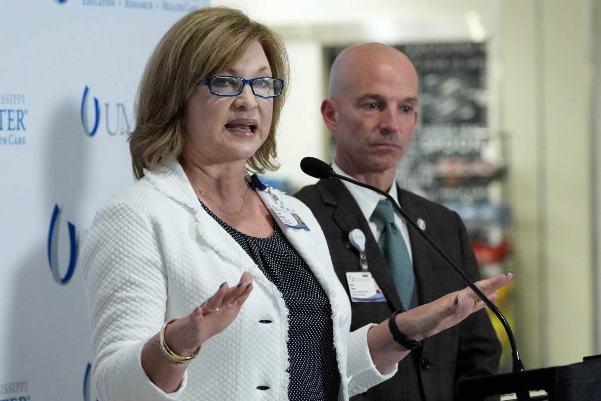 A woman with a white coat speaking. A man wearing a suit stands behind her.