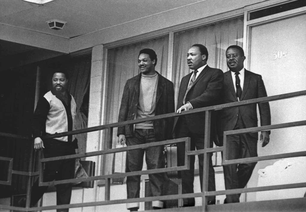 An old black and white photo of four men, including Jesse Jackson and Martin Luther King Jr. standing on the balcony outside of a hotel room