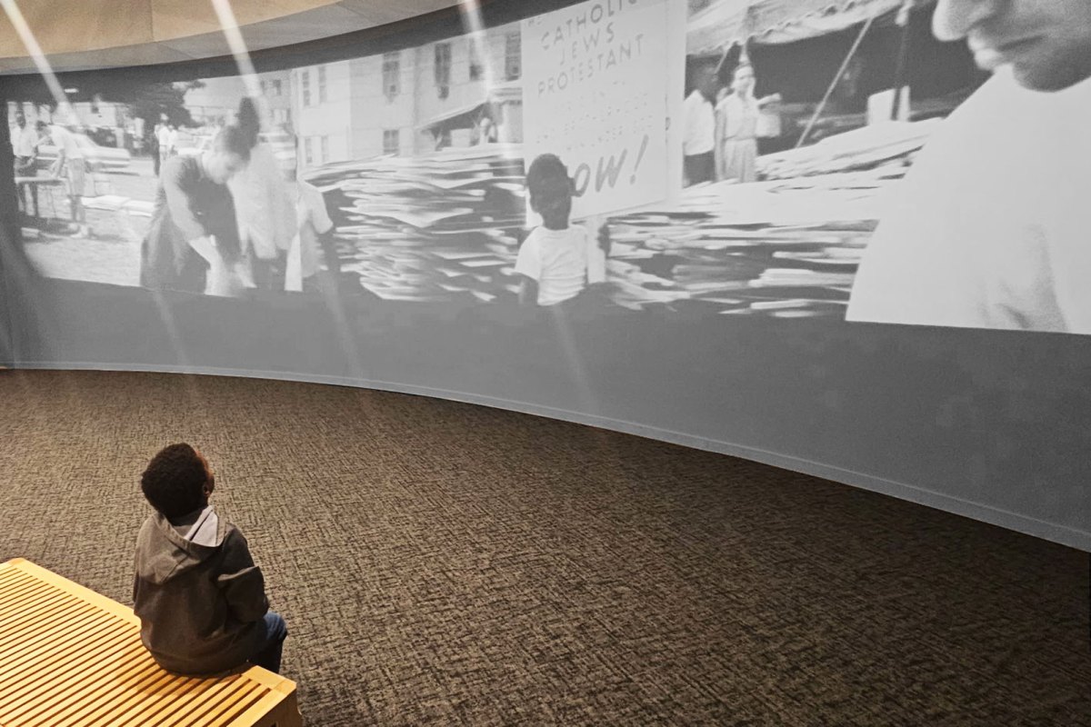 A young boy sits on a museum bench and looks up at a large projected exhibit about the civil rights
