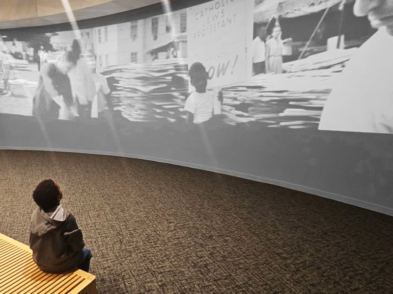 A young boy sits on a museum bench and looks up at a large projected exhibit about the civil rights