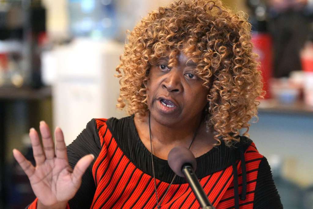 Omeria Scott, wearing black and red, speaking at a mic with one hand gesturing before her