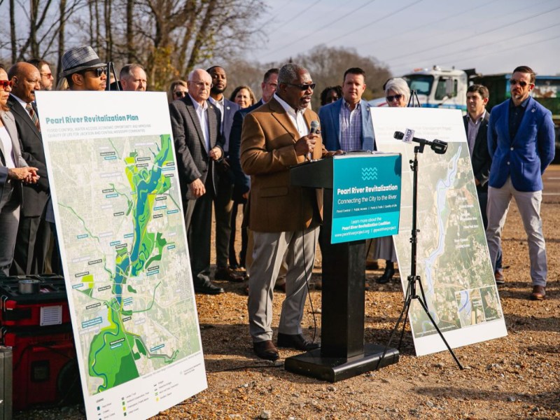 photo of a man speaking with people behind him and maps on either side