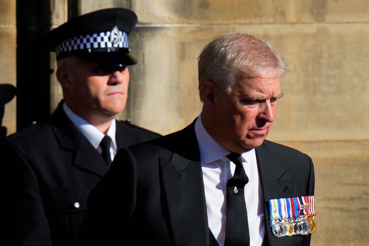 Prince Andrew wearing a suit and several medals walking forward. A security guard walks behind him, and in front of him, another guard stands holding a pole.
