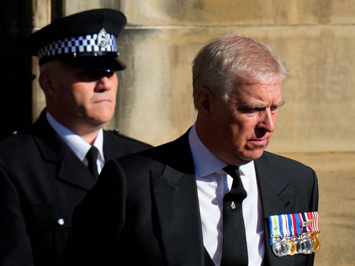 Prince Andrew wearing a suit and several medals walking forward. A security guard walks behind him, and in front of him, another guard stands holding a pole.