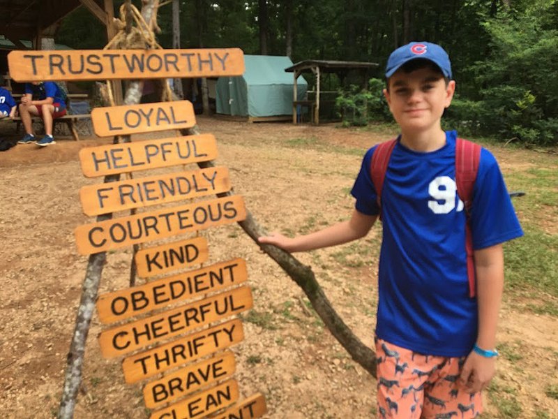 A young boy wearing a blue shirt and baseball cap. He is standing next to a sign featuring several words describing scout values.