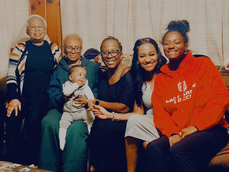 Five women of different generations sit on a couch together, one holding a small baby