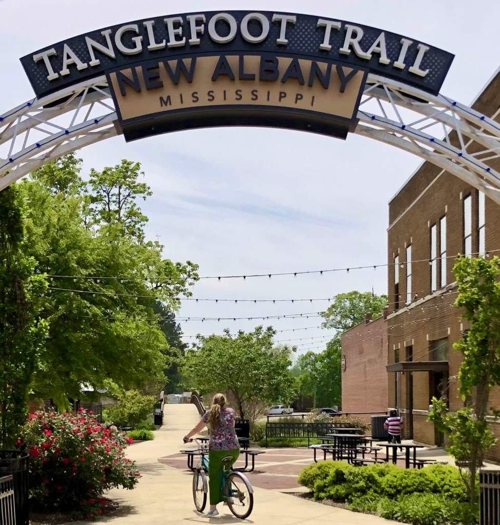 A sign leading to a park that reads, “Tanglefoot Trail.” Beneath it is another sign that reads, “New Albany, Mississippi.”
