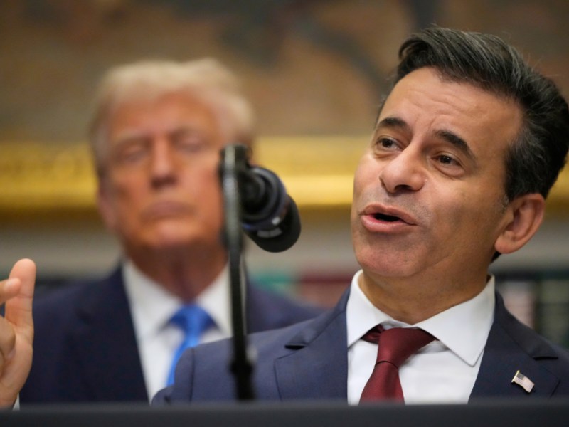 A man wearing a suit speaking into a microphone. Donald Trump is behind him.