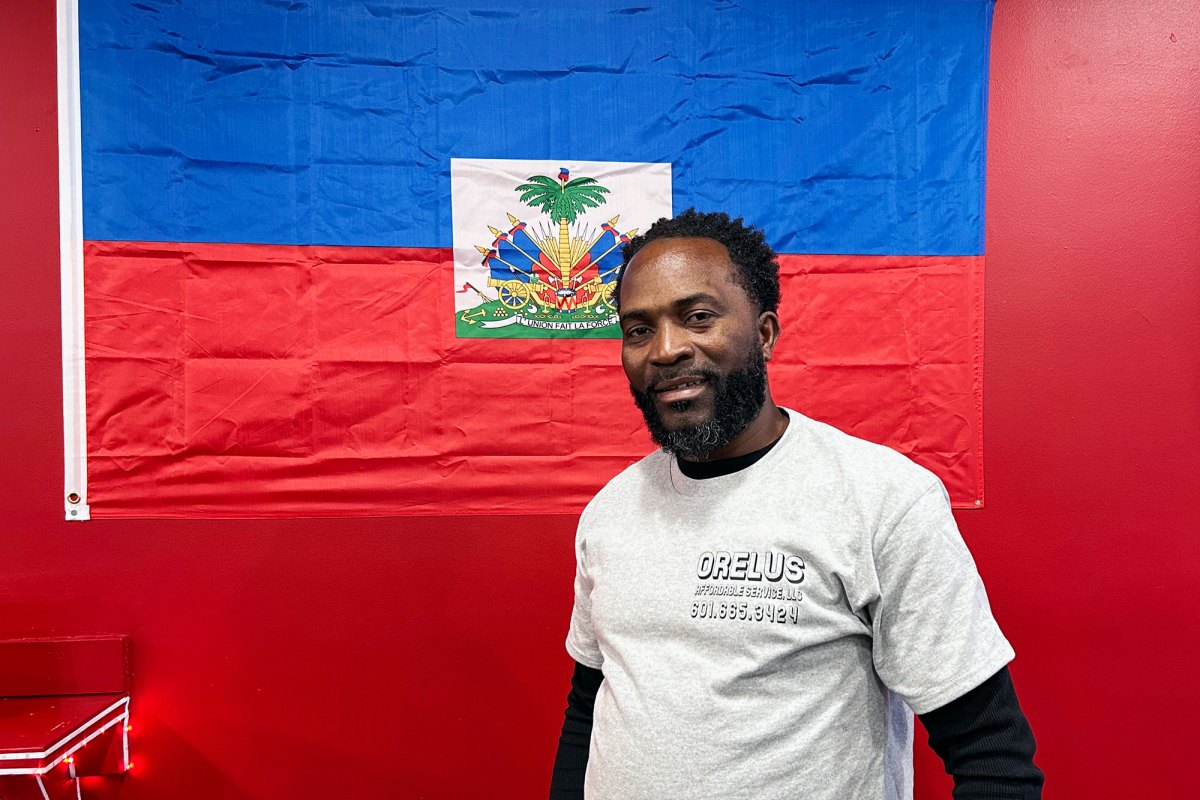 A man stands in front of a red wall with the flag of Haiti hanging
